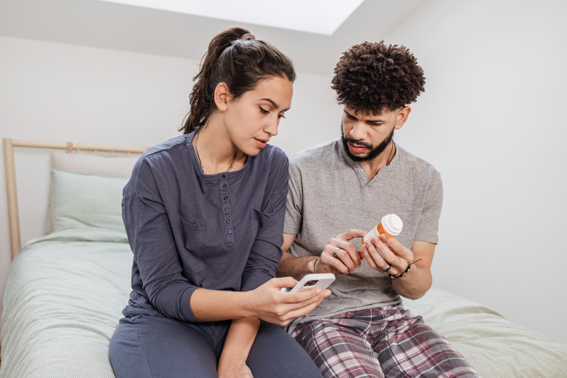 Couple sitting on a bed, reading a prescription bottle while checking a phone