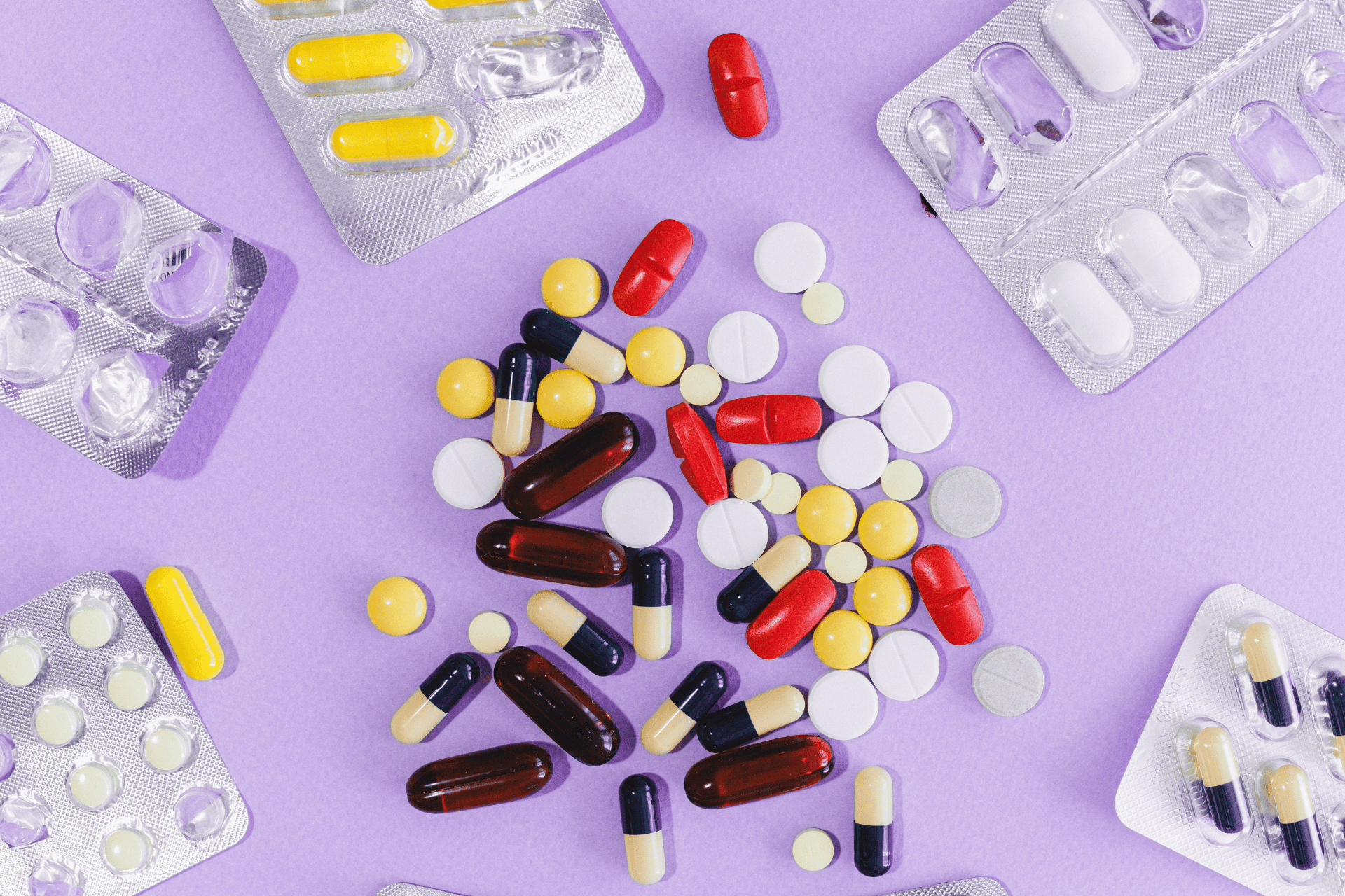 Assorted painkillers and medications, including tablets and capsules of various colors, scattered on a purple background with blister packs nearby.