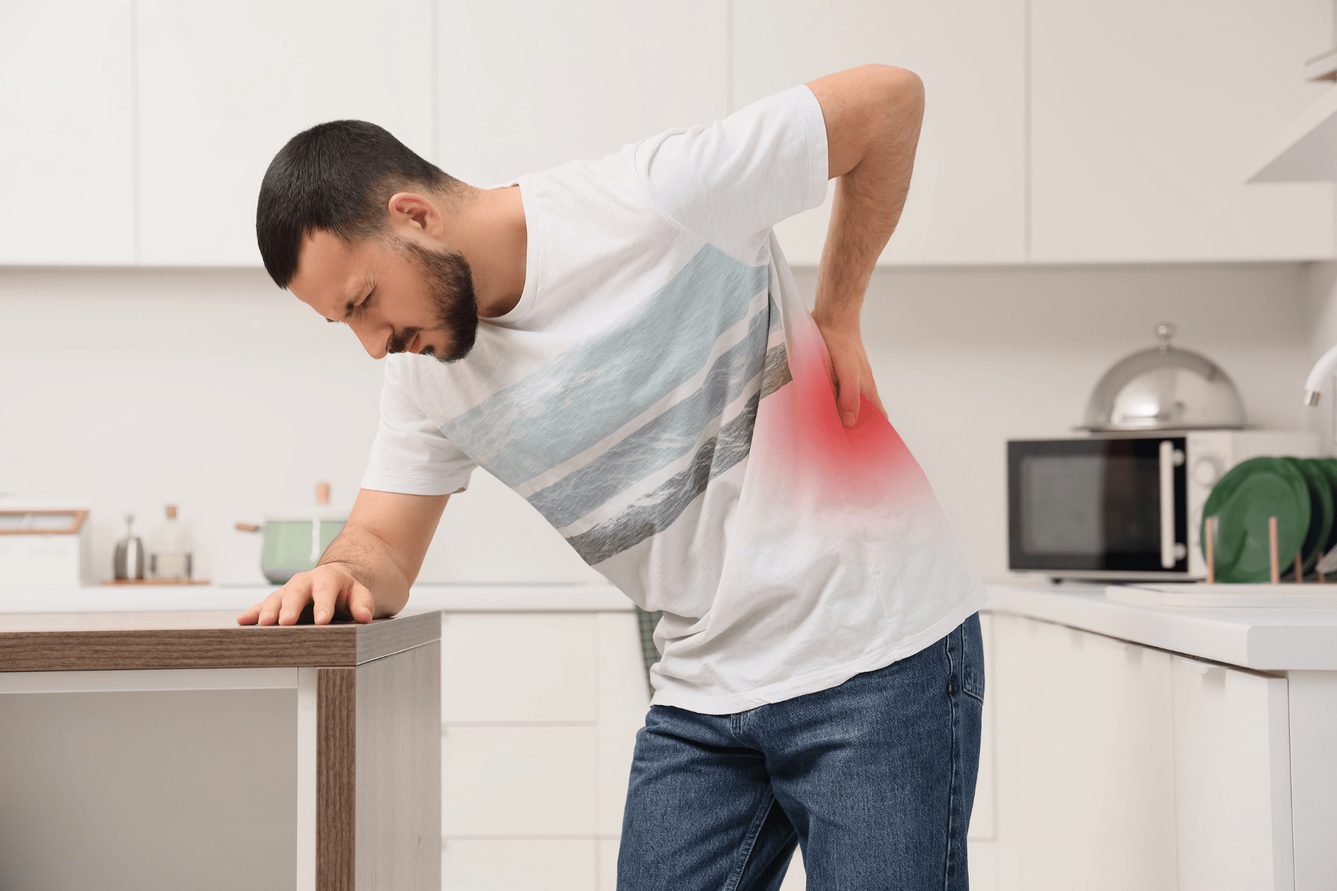 A man in a kitchen braces himself on a counter while holding his lower back, with a red highlighted area indicating acute back pain.
