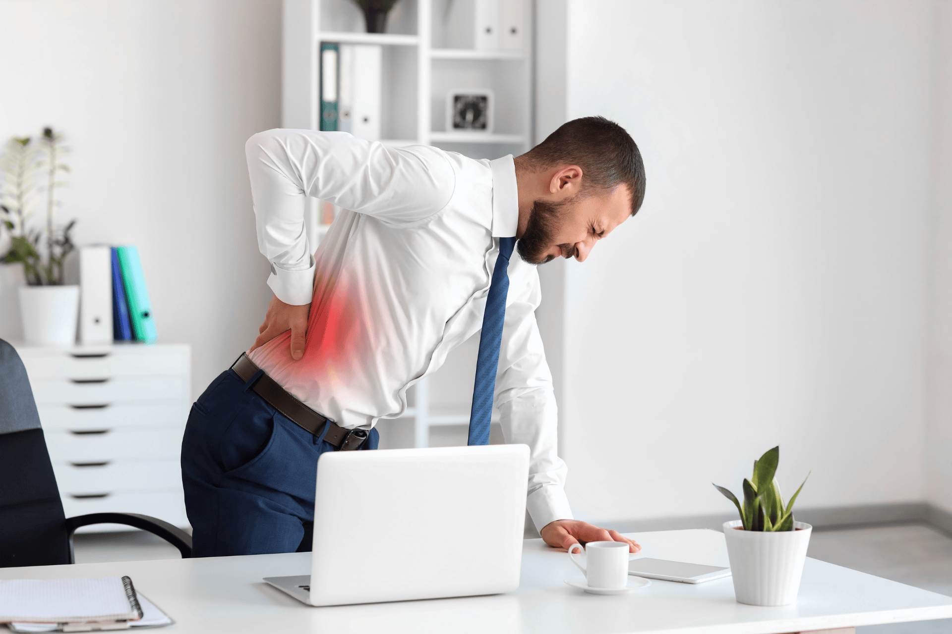 A man in an office leans over a desk while holding his lower back, with a red highlighted area indicating back pain.