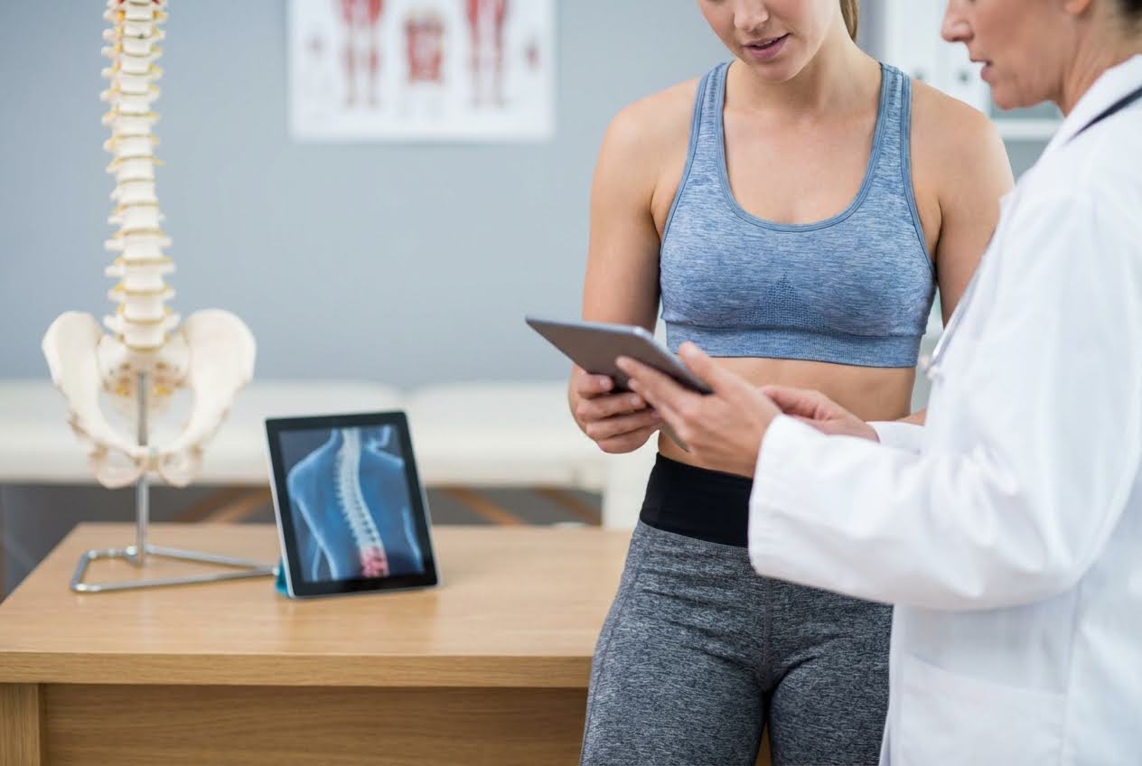 Doctor and patient discussing a spinal X-ray on a tablet, with a spine model on a table