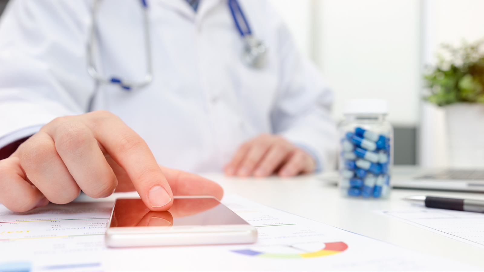 A doctor in a white coat uses a smartphone at a desk with documents, pills, and a laptop, representing digital healthcare