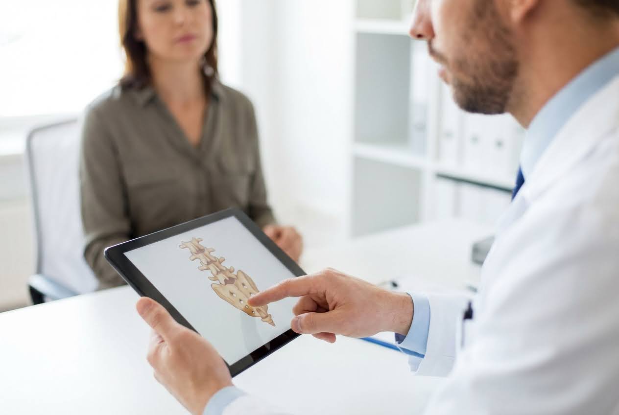 Doctor in white coat points to a tailbone diagram on a tablet for a female patient in a bright office