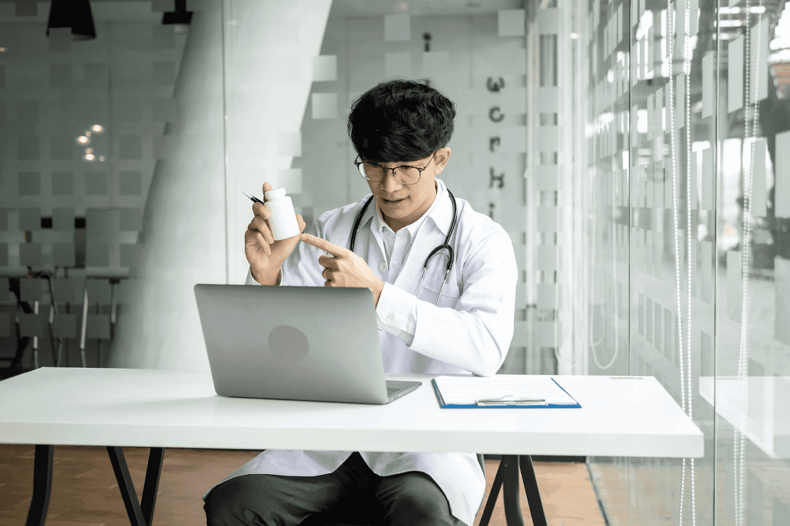 Doctor pointing to a pill bottle while on a video call at a desk with a laptop.