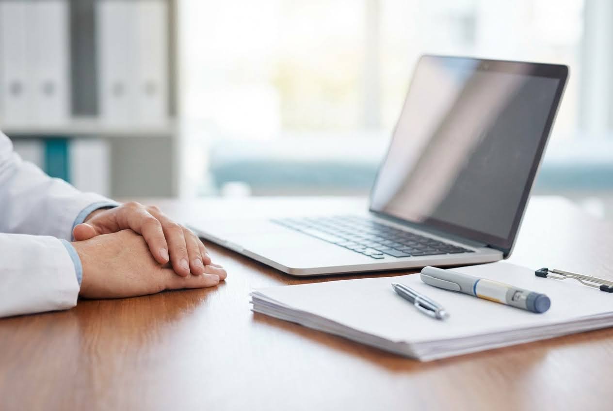 Doctor's hands on a wooden desk with a laptop, pen, and an insulin pen on a stack of papers