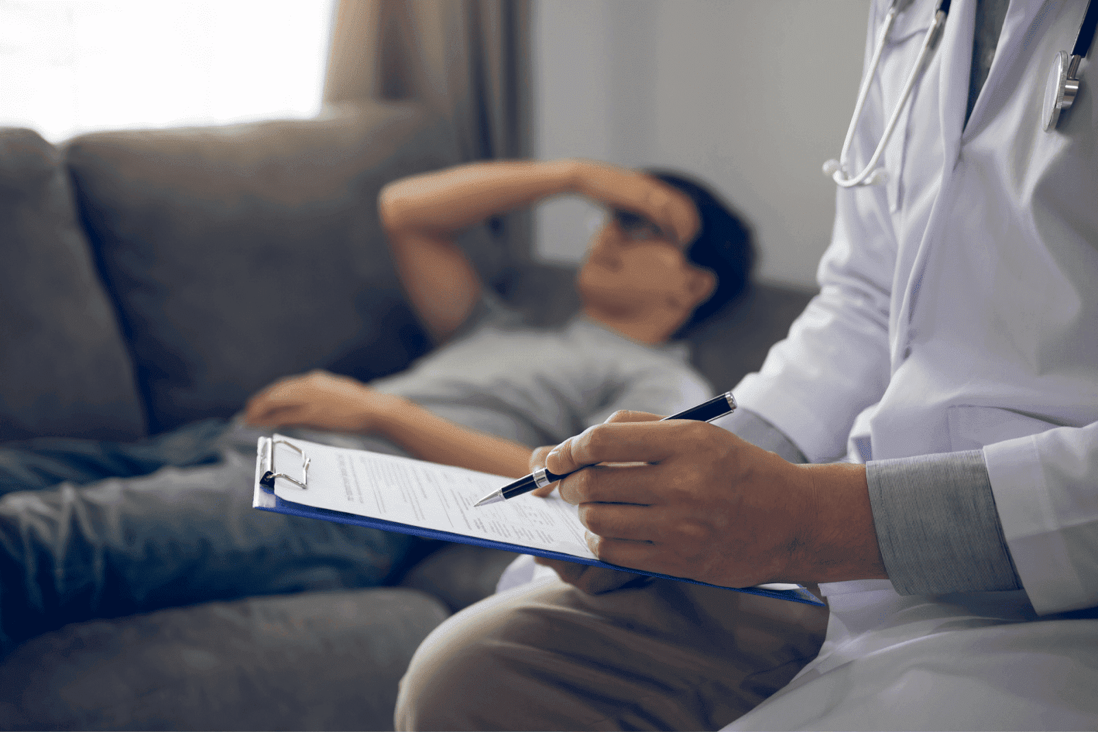 Doctor taking notes while patient lies on couch, hand on forehead.