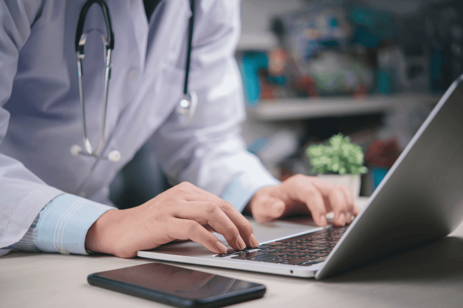 Doctor using a laptop with a stethoscope around the neck and a phone on the desk.