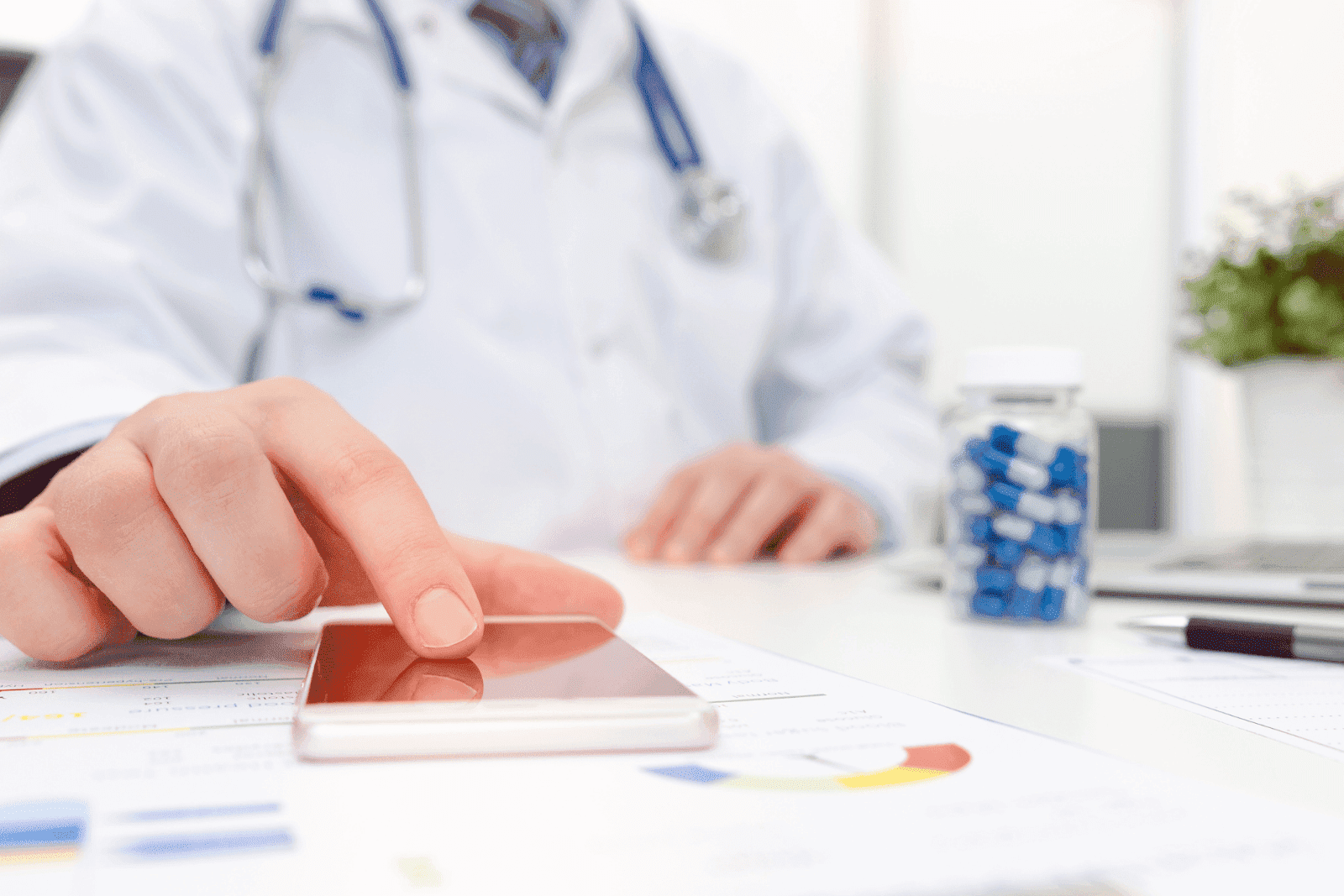 A doctor in a white coat with a stethoscope around their neck uses a smartphone at a desk covered with medical charts, a bottle of blue and white capsules, and a laptop in the background.
