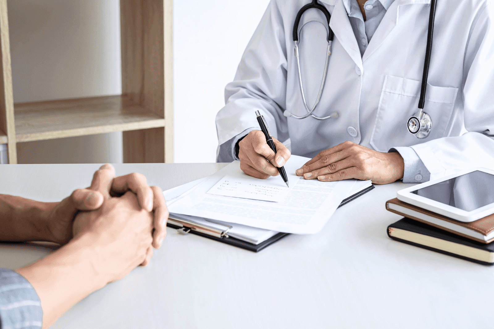Doctor writing notes on a clipboard while speaking with a patient during a medical consultation at a desk.