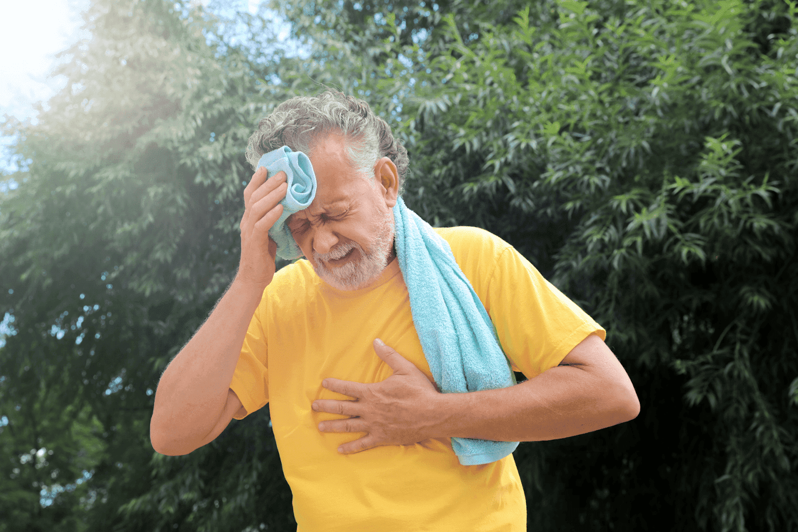 Elderly man in a yellow shirt outdoors, sweating and clutching his chest with a pained expression