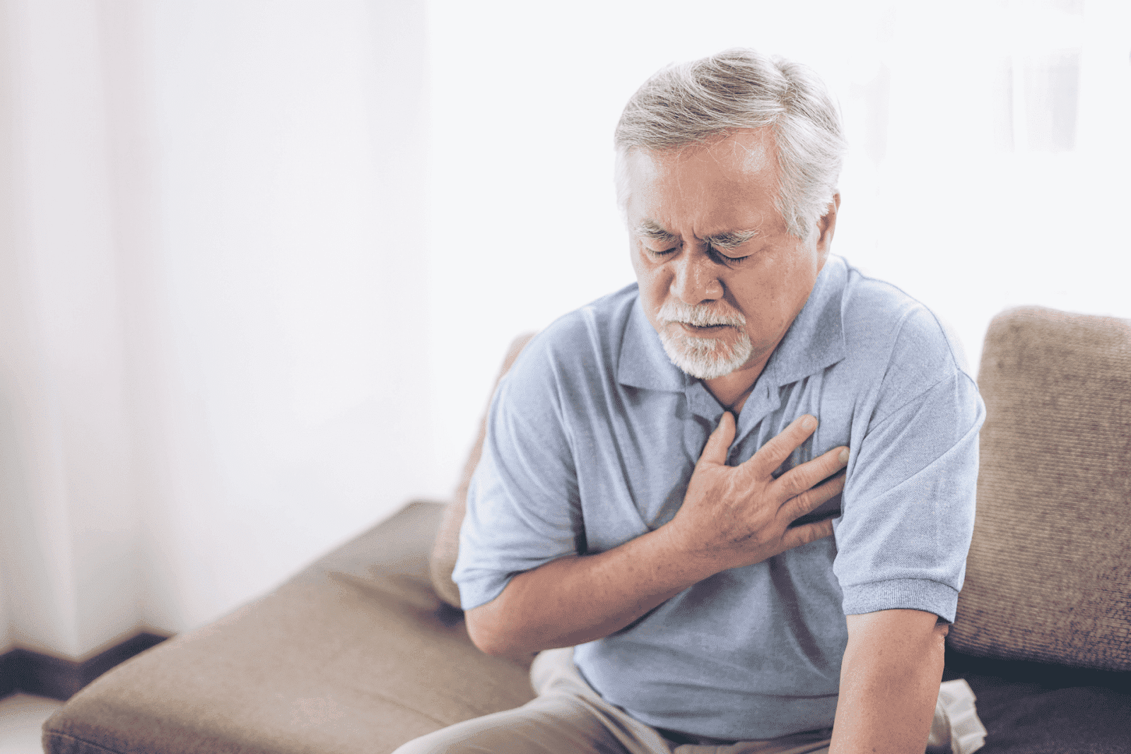 Elderly man sitting on a couch, clutching his chest in pain, appearing to experience chest discomfort or a possible heart issue.