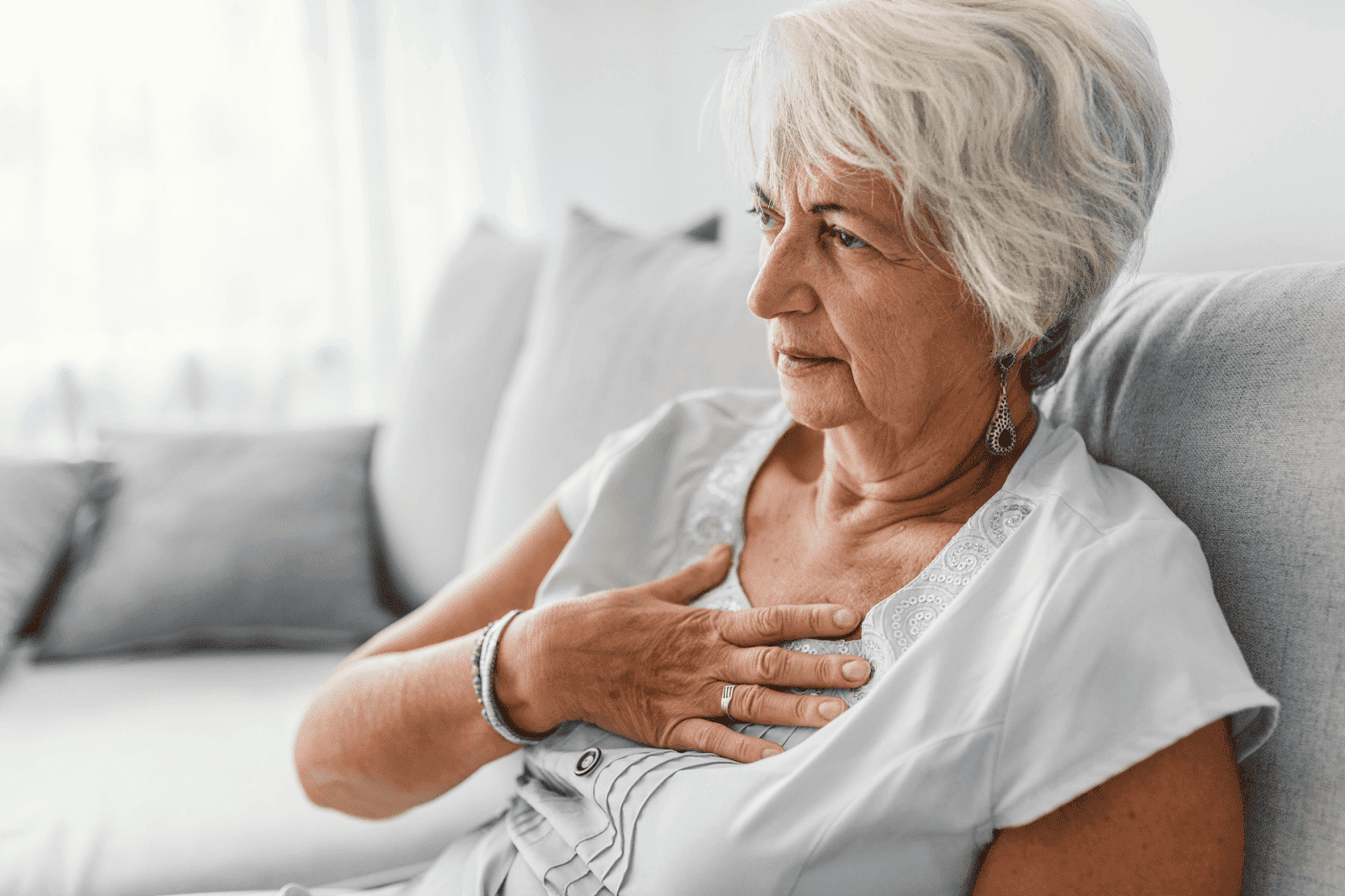 Elderly woman sitting on a couch with her hand on her chest, appearing uncomfortable.