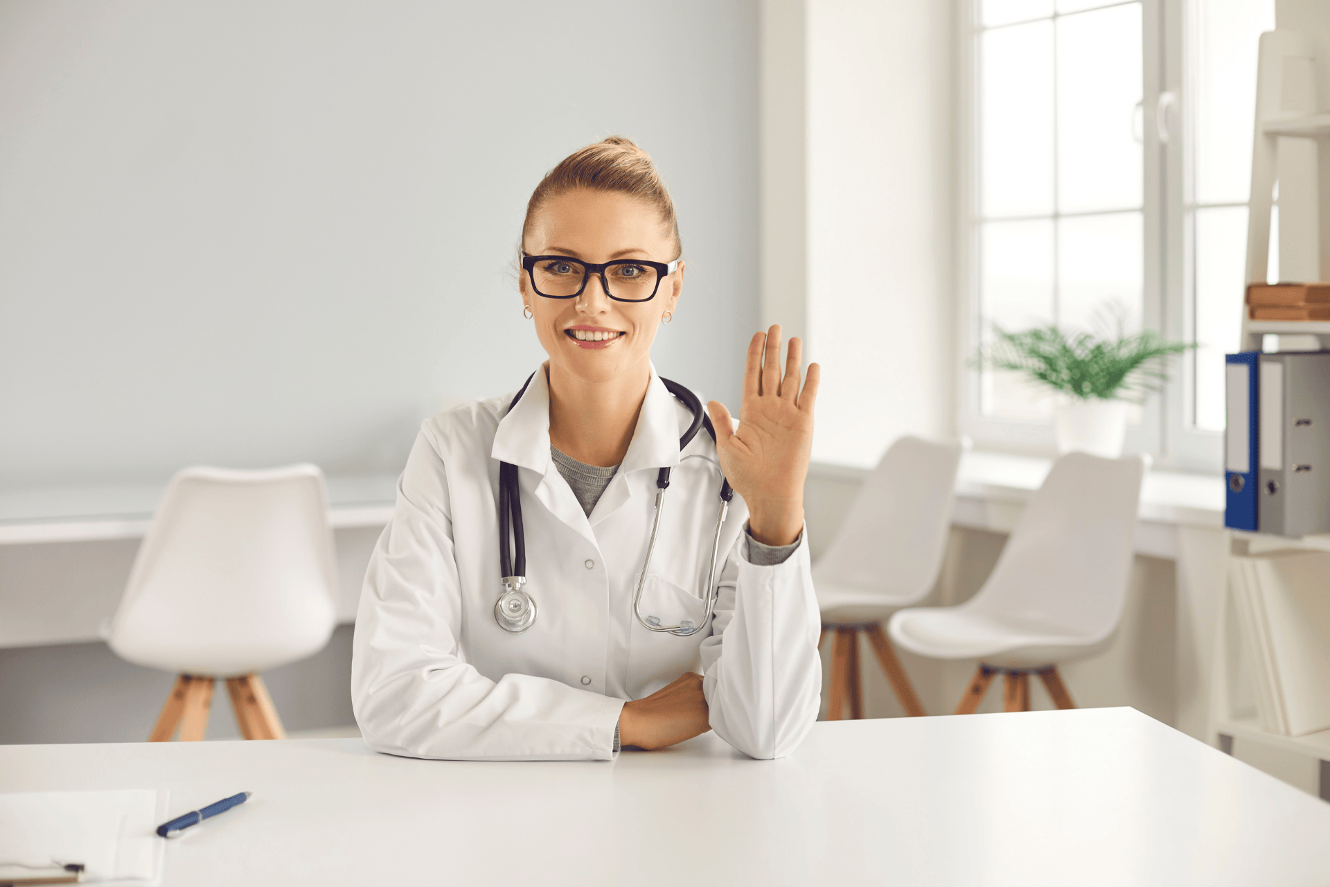Female doctor in a white coat smiling and waving during a consultation.