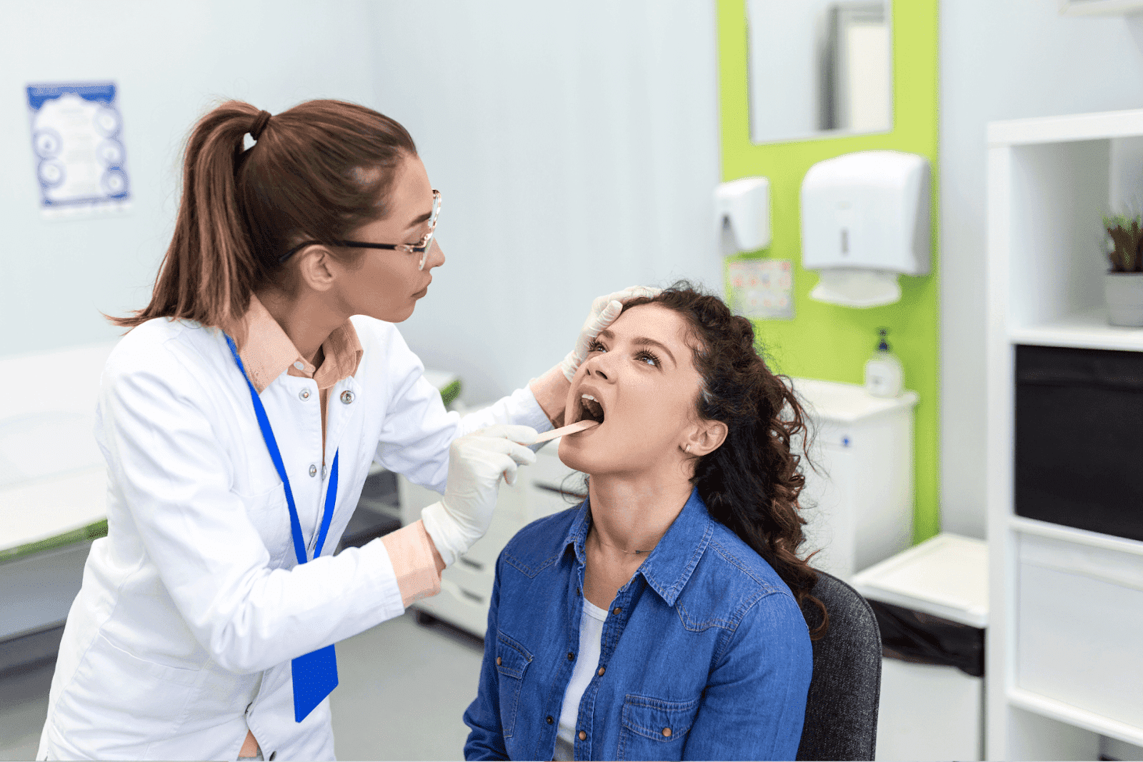Female doctor in a white coat using a tongue depressor to examine the throat of a seated woman in a medical clinic