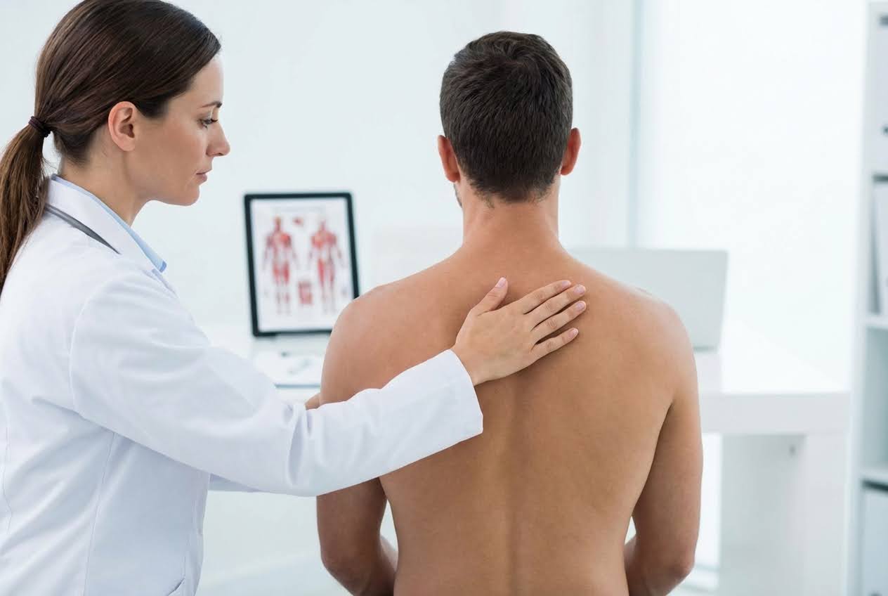 Female doctor in white coat examining a shirtless male patient's upper back, with an anatomical chart in the background.