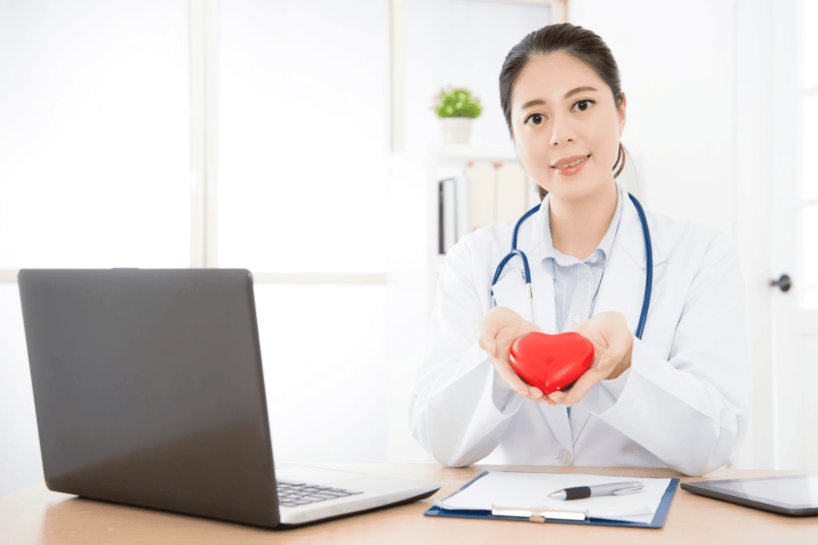 Female doctor smiling while holding a red heart model in her hands at a desk with a laptop