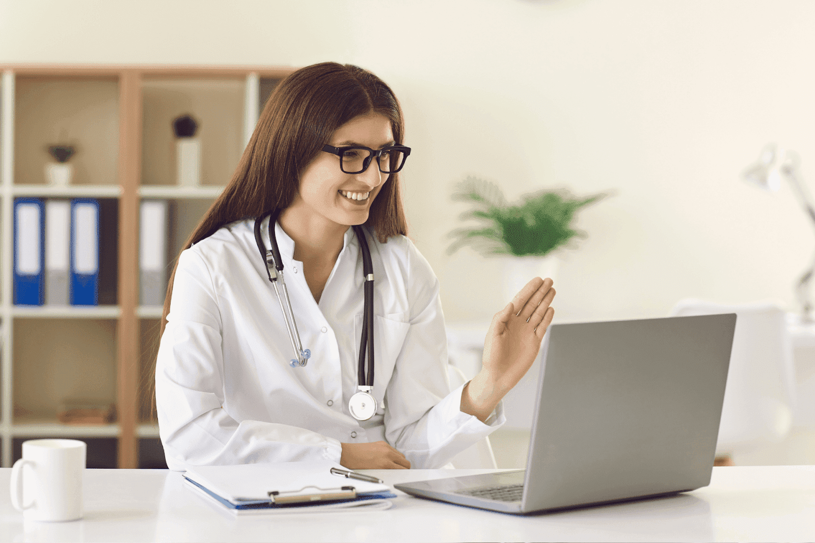 Female doctor waving during a video call on her laptop.