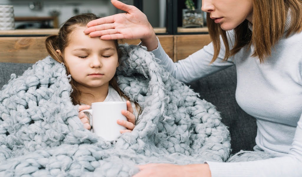 Feverish girl holding a hot drink under a blanket, mother comforting her