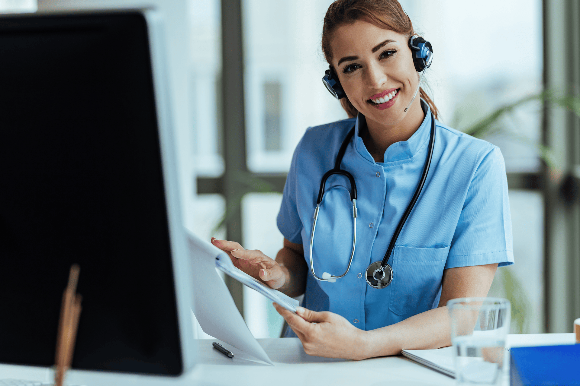 Smiling nurse with a headset and stethoscope working at a computer.