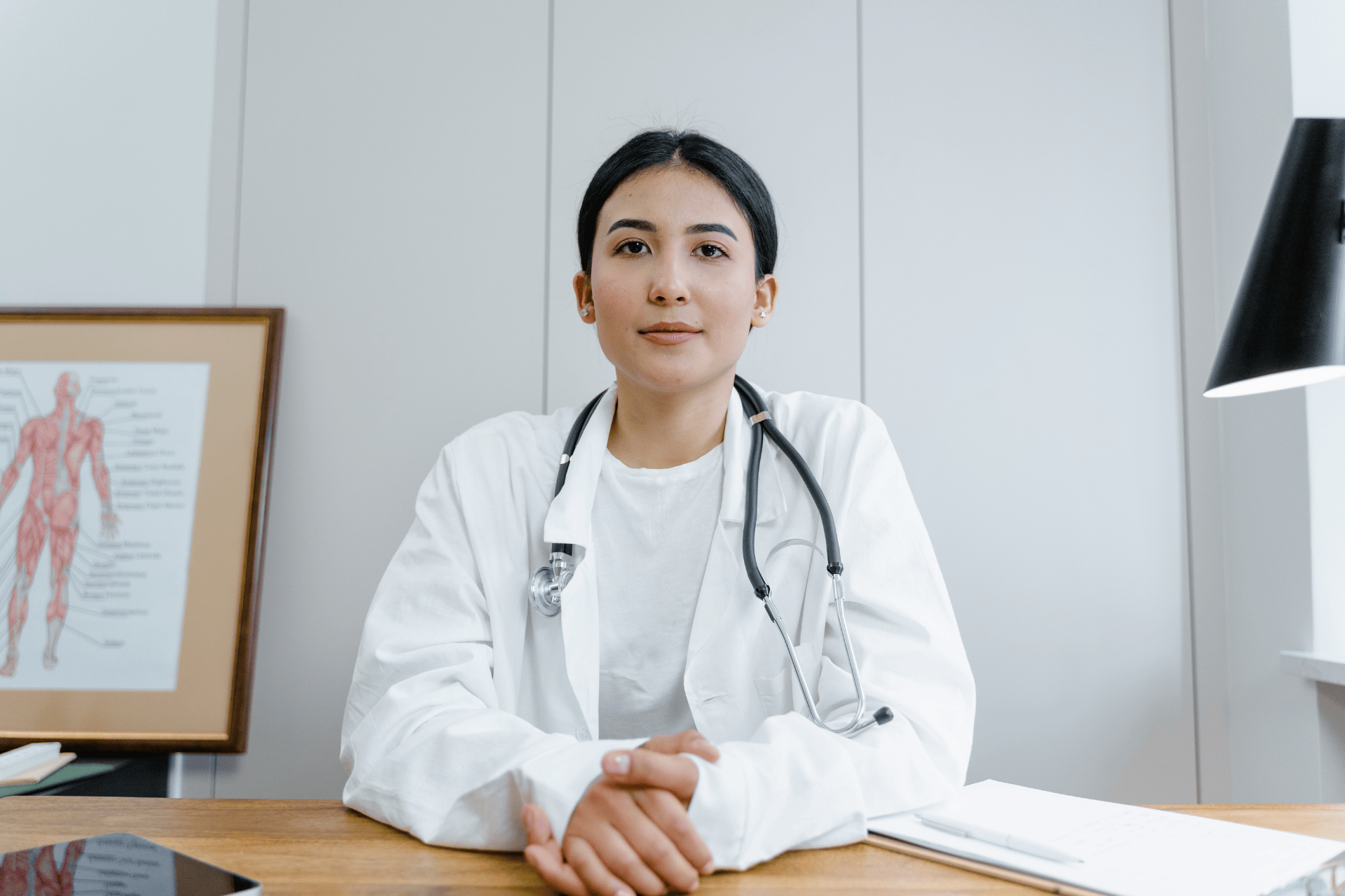 Doctor sitting at a desk with a stethoscope, looking directly at the camera.