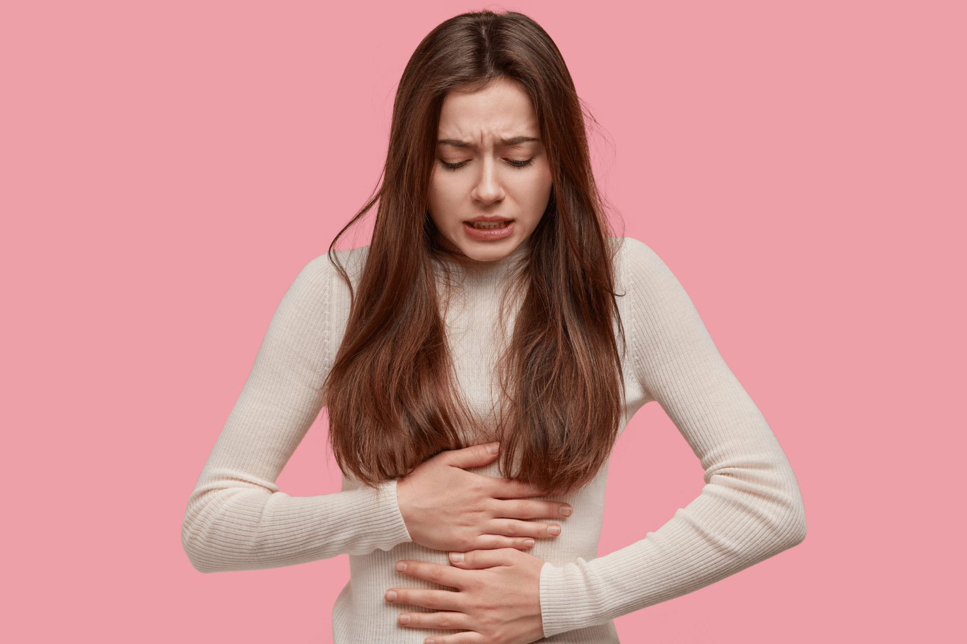 Young woman holding her stomach in discomfort against a pink background.