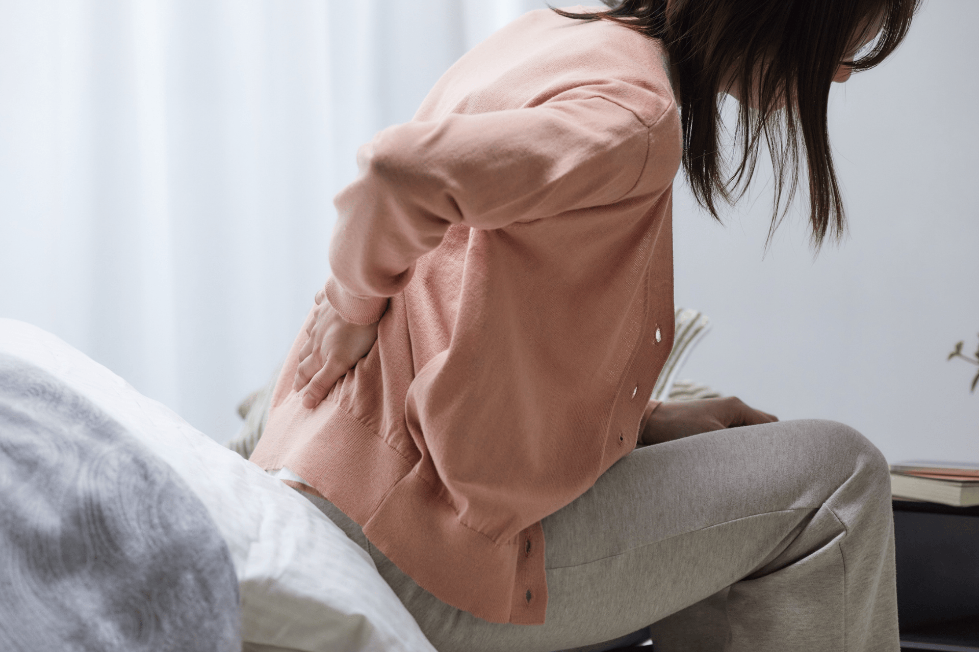 A person sitting on the edge of a bed, holding their lower back in apparent pain or discomfort, possibly indicating back pain.