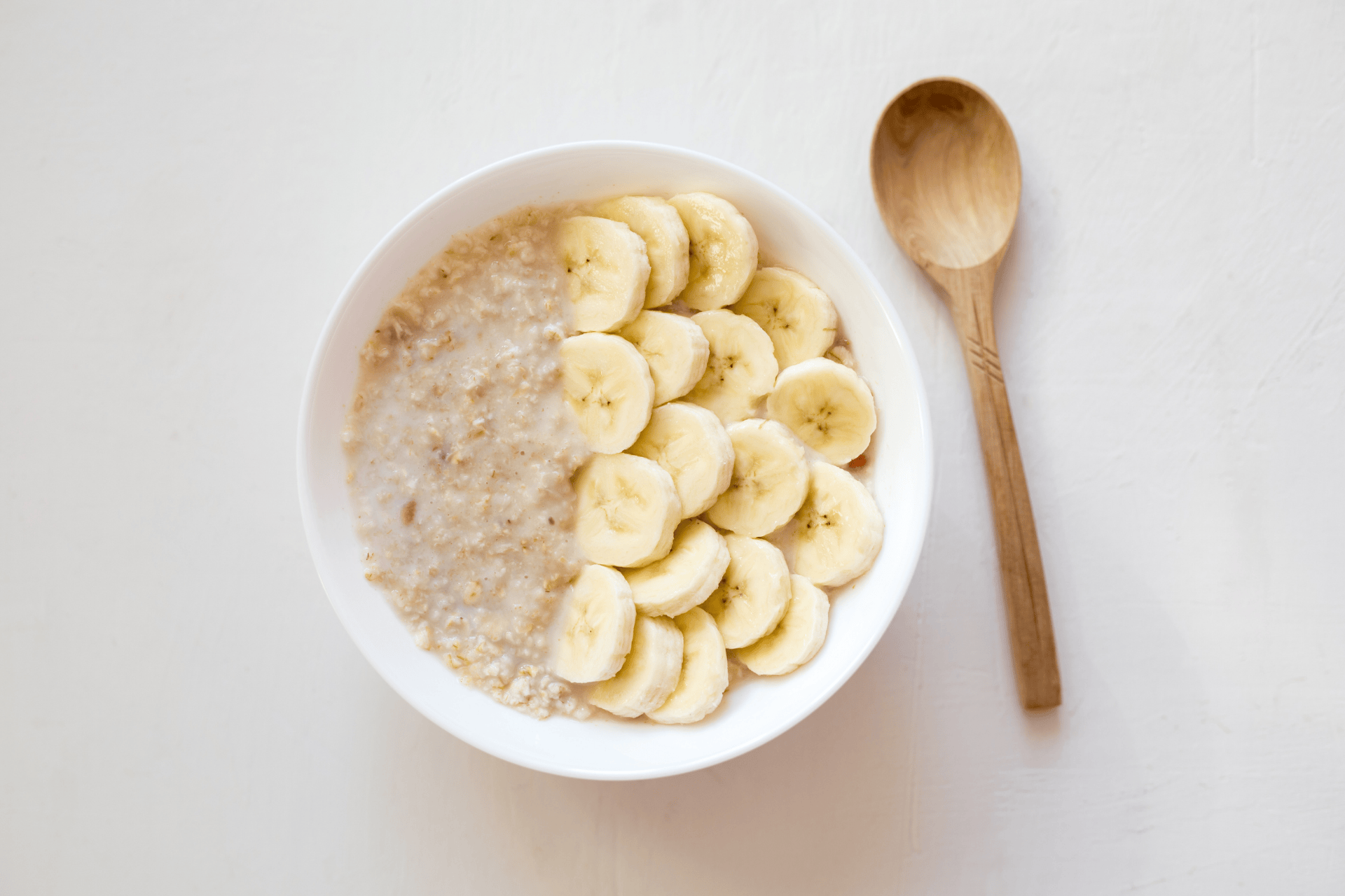 Bowl of oatmeal topped with sliced bananas beside a wooden spoon.