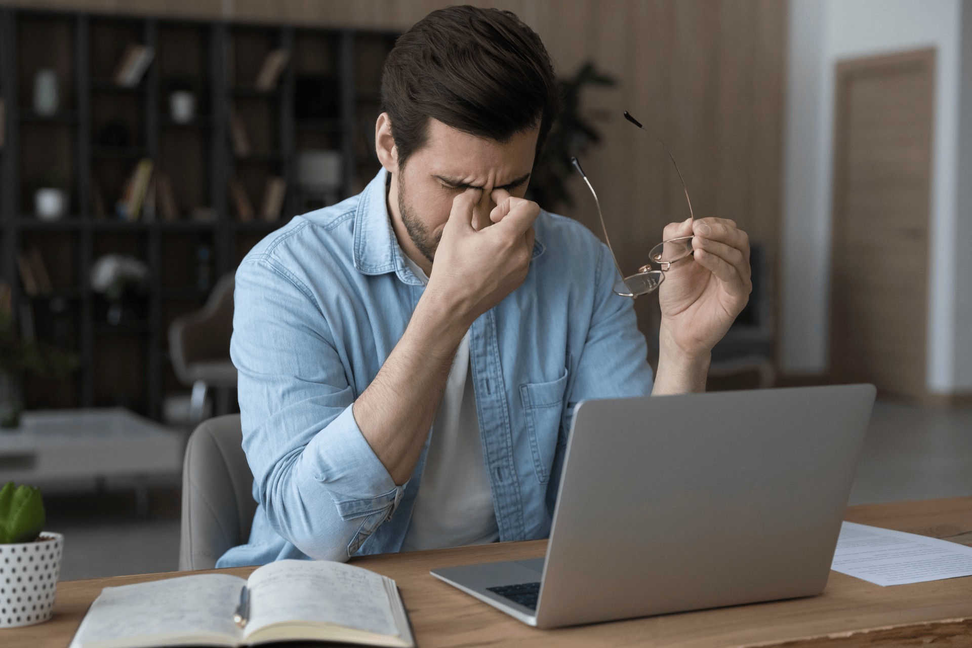 A Man sitting at a desk, rubbing his eyes from fatigue while using a laptop