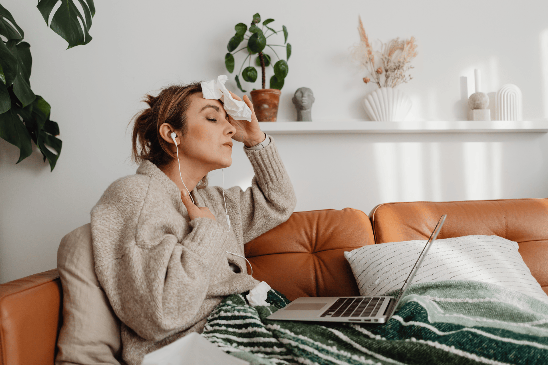 Sick woman with earphones holding a tissue to her forehead during an online consultation.