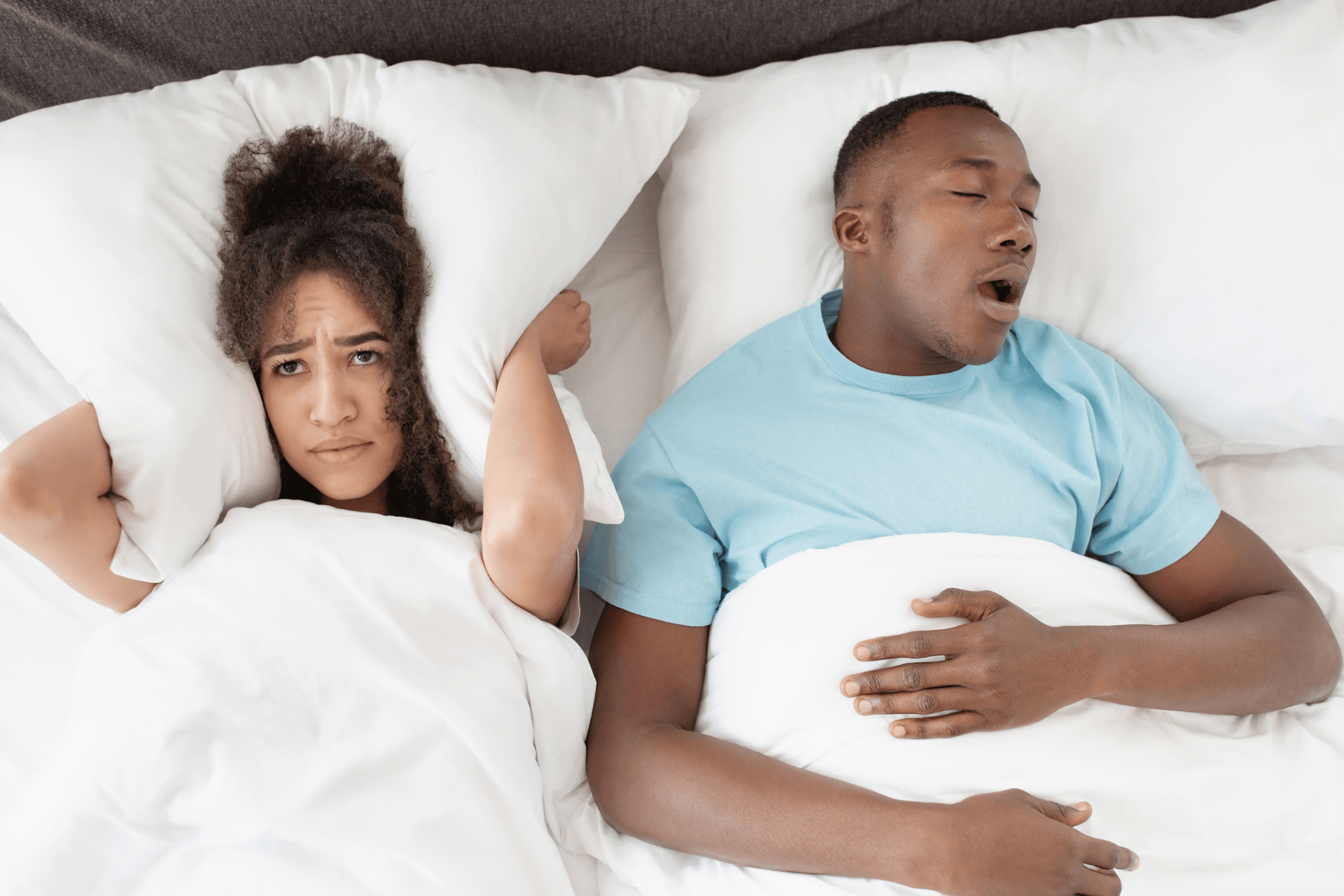 Woman covering her ears with a pillow while a man next to her snores.