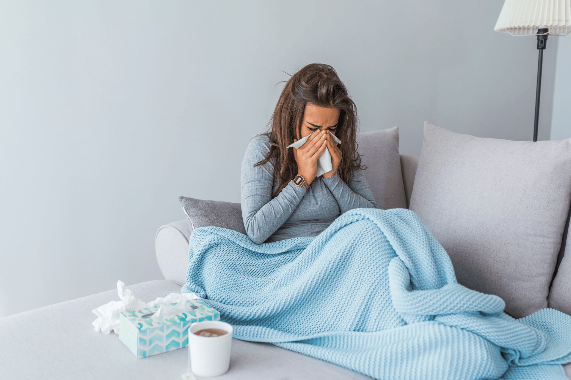 Woman sitting on couch blowing her nose under a blue blanket.