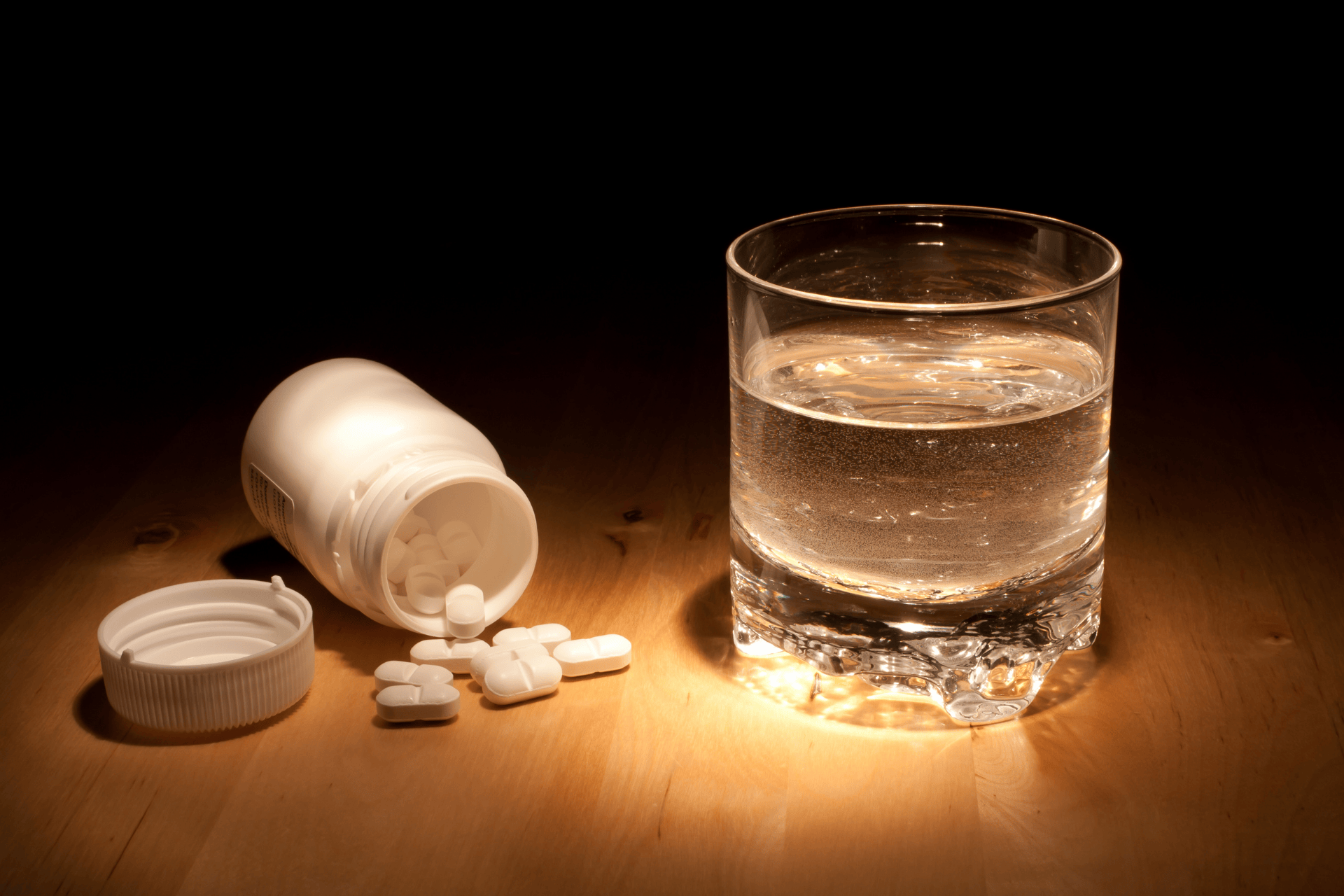Glass of water and spilled white pills from an open bottle on a wooden table.