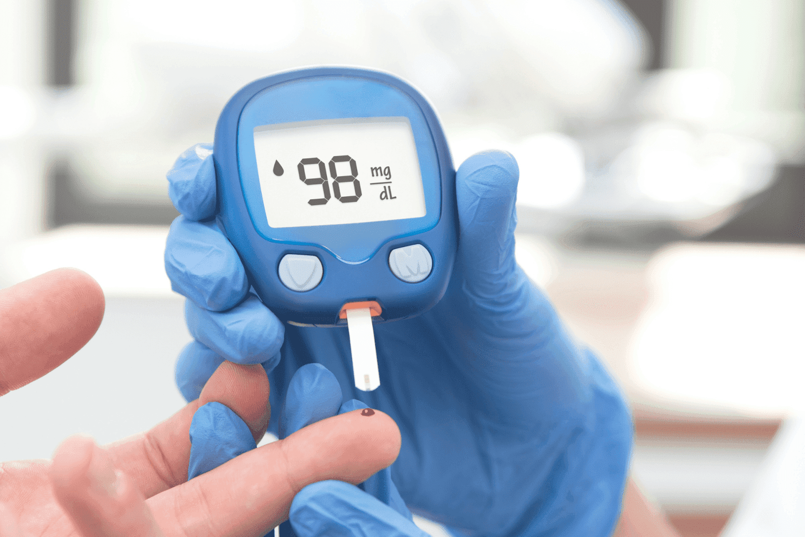 Gloved hand holding a glucose meter showing a reading of 98 mg/dL during a blood sugar test