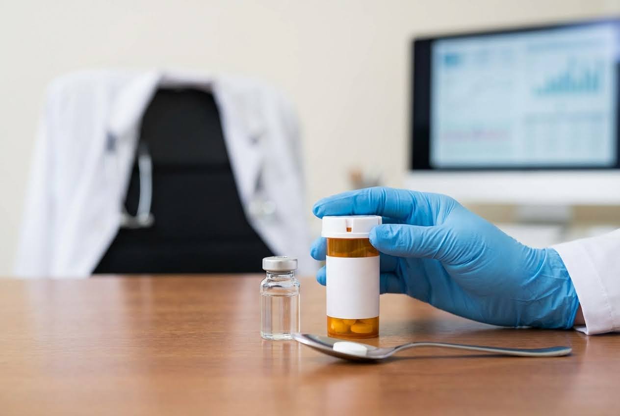 Gloved hand holding a pill bottle, with a vial, spoon, and pills on a wooden desk, a doctor's coat, and a monitor in the background