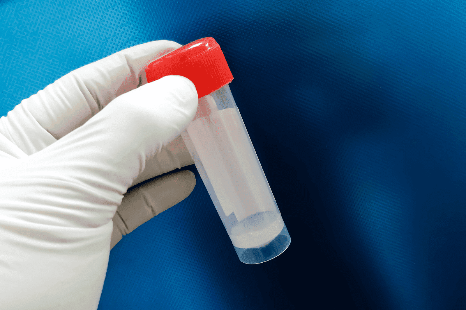 Gloved hand holding a sealed container with a sperm sample against a blue background
