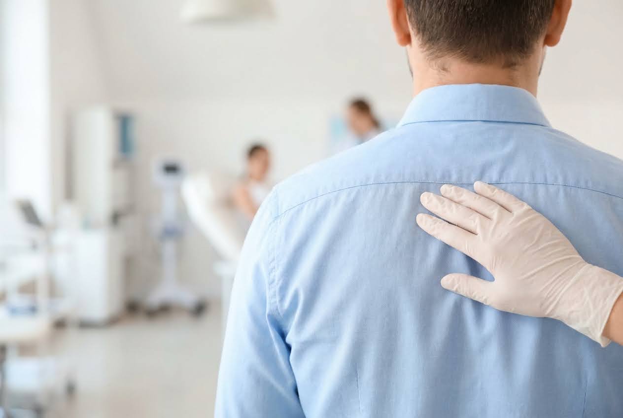 Gloved hand touching a man's blue-shirted upper back, with a blurred clinic room in the background