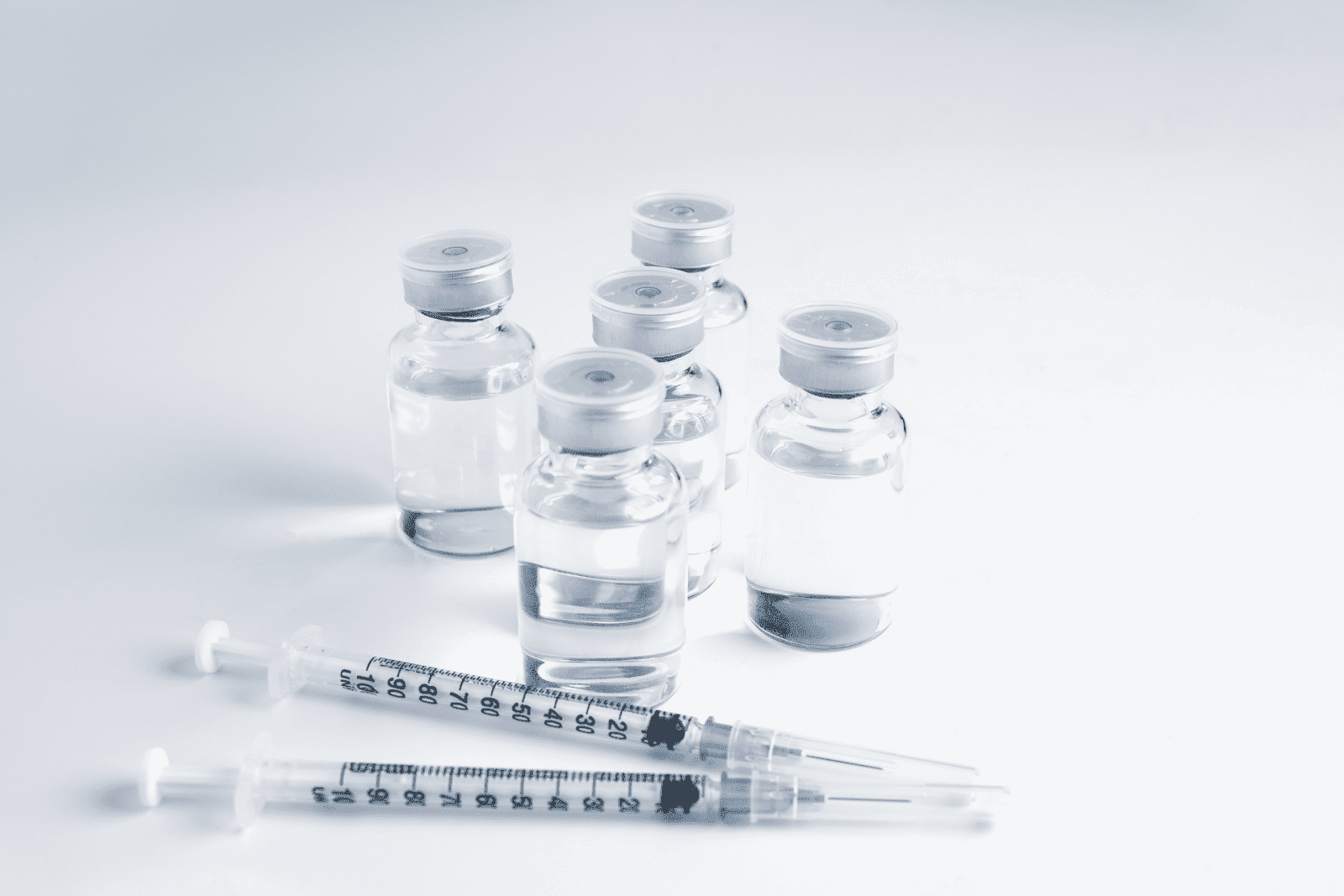 Group of medical vials with clear liquid and two syringes on a white surface.