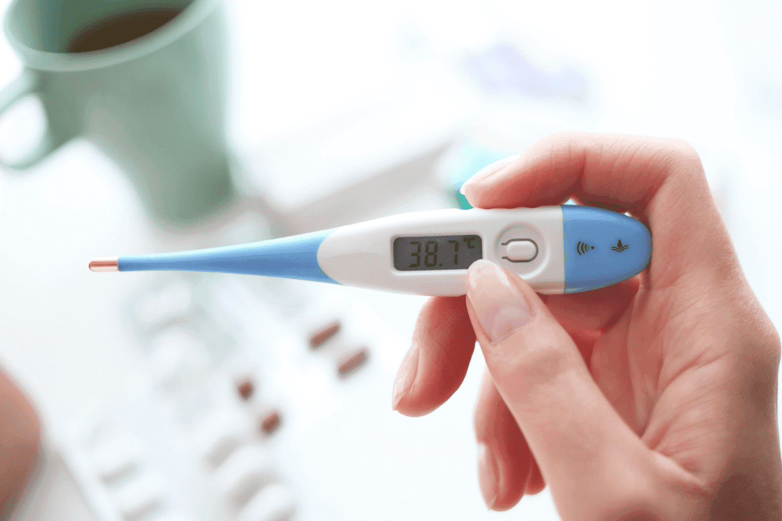 Hand holding digital thermometer showing 38.7°C fever.
