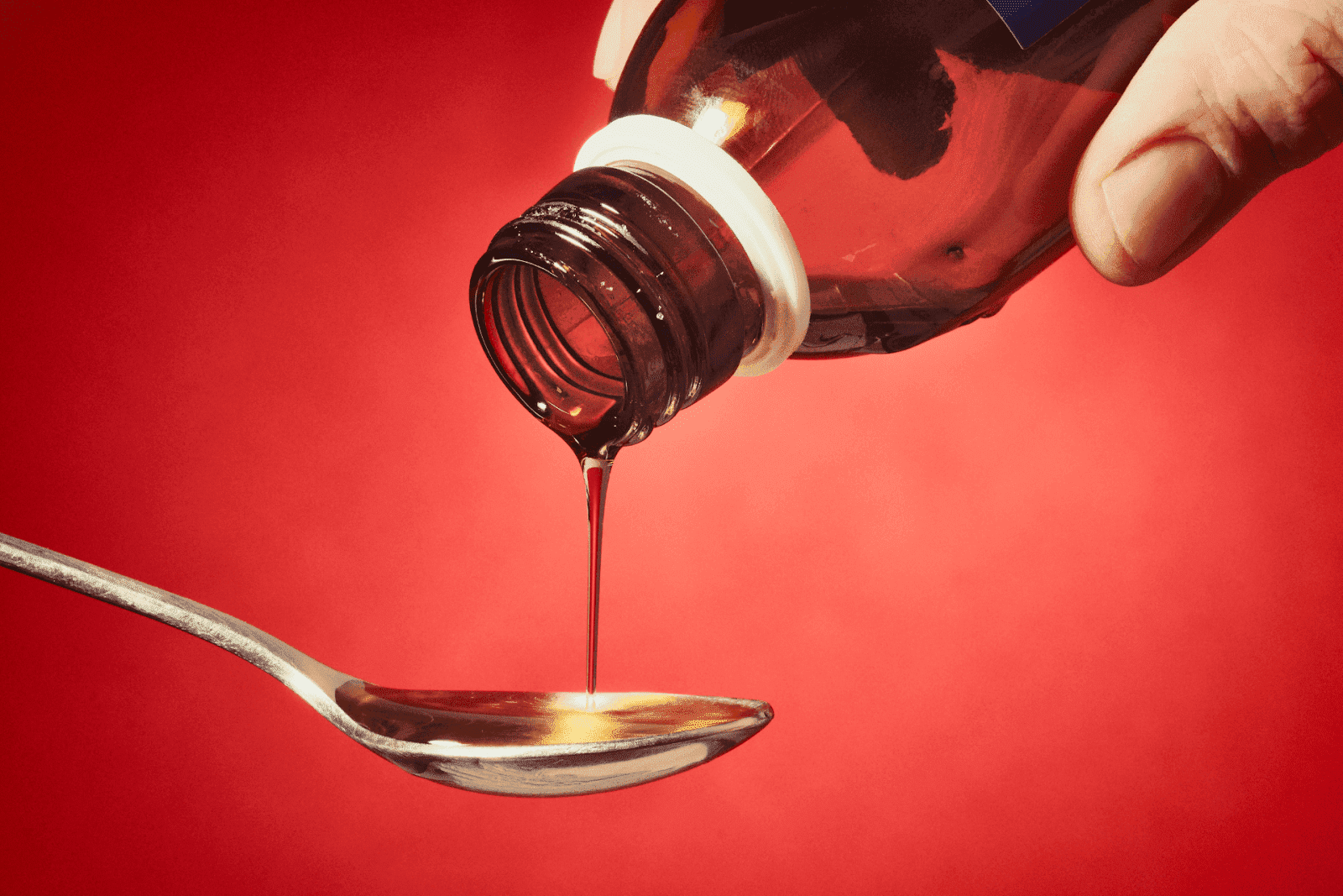 Liquid medicine being poured from a bottle into a spoon against a red background.