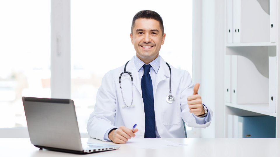 Healthcare professional at desk with stethoscope and laptop, giving a thumbs-up