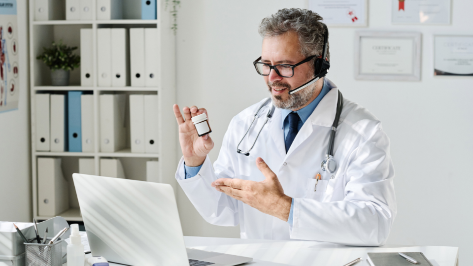 A doctor wearing a headset and white coat holding a medication bottle during an online consultation