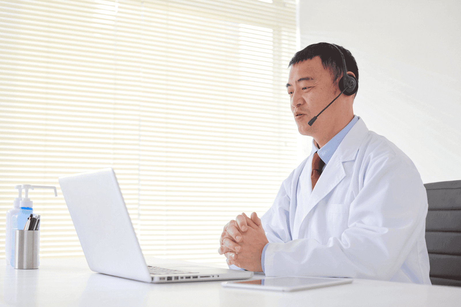 Male doctor in a white coat with a headset conducting a telehealth visit on a laptop
