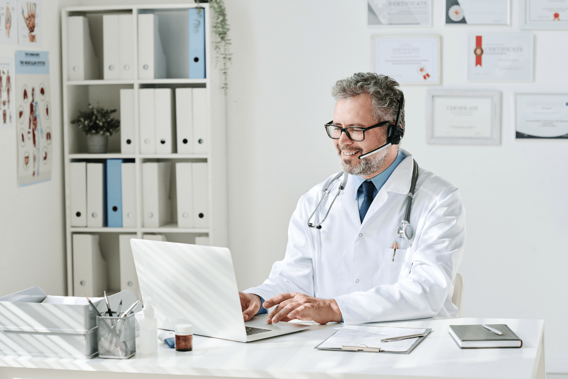 Male doctor wearing a headset smiles while working on a laptop in his office.