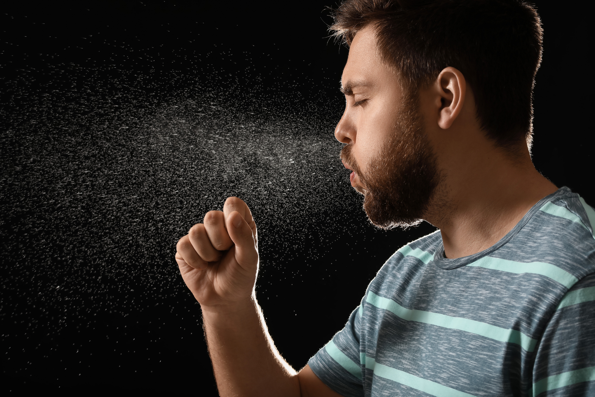 Man coughing into his fist with visible respiratory droplets in the air against a dark background