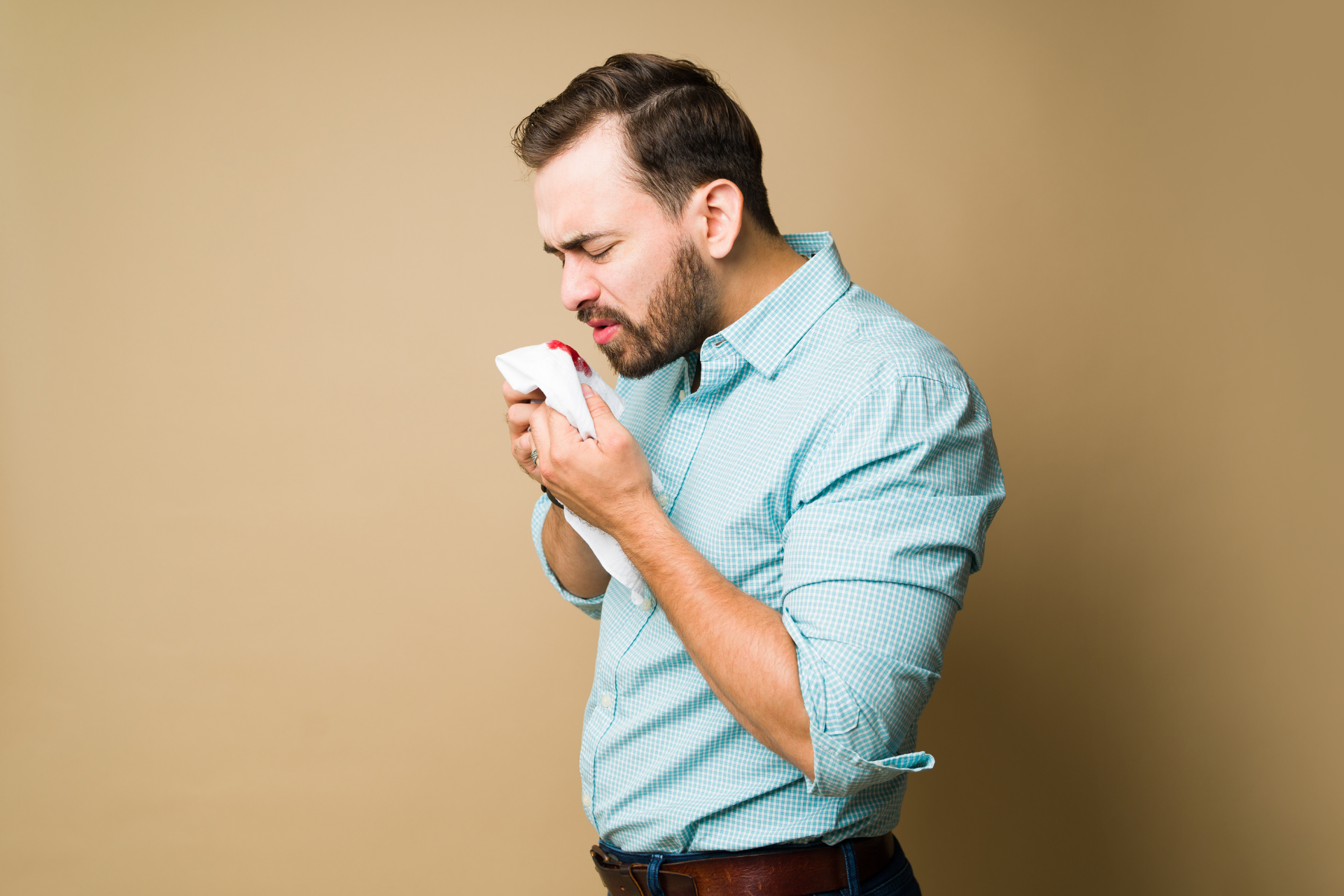Man holding a tissue to his nose with visible blood, appearing distressed from a nosebleed indoors