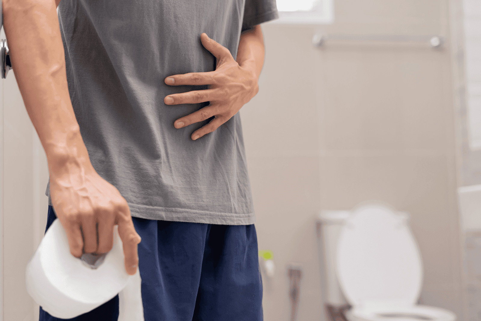 Man holding his stomach in pain while standing in a bathroom with toilet paper in hand