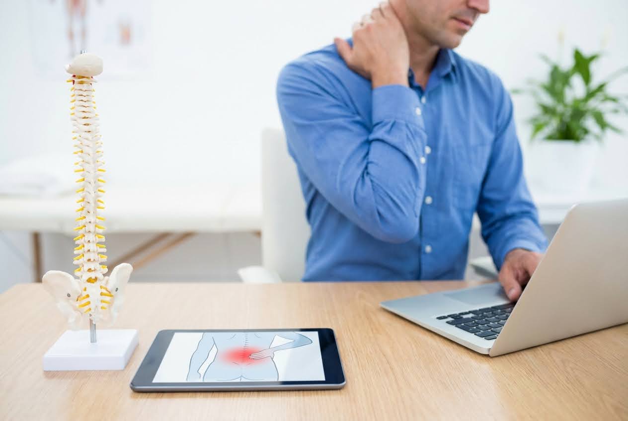 Man in blue shirt rubbing his neck, with a spine model and tablet showing back pain on a desk.