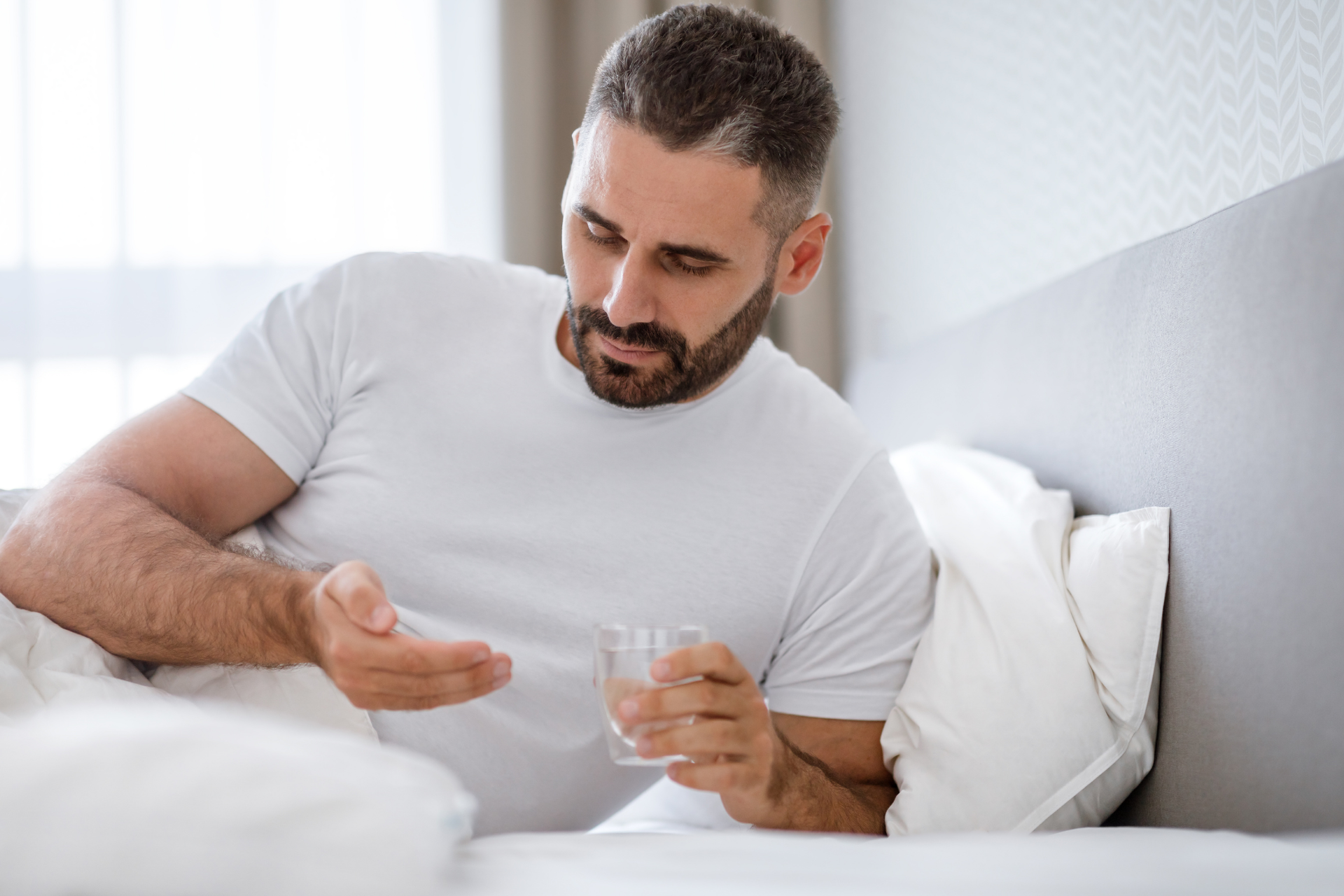 Man lying in bed holding a glass of water and examining medication in his hand, appearing unwell or recovering from illness