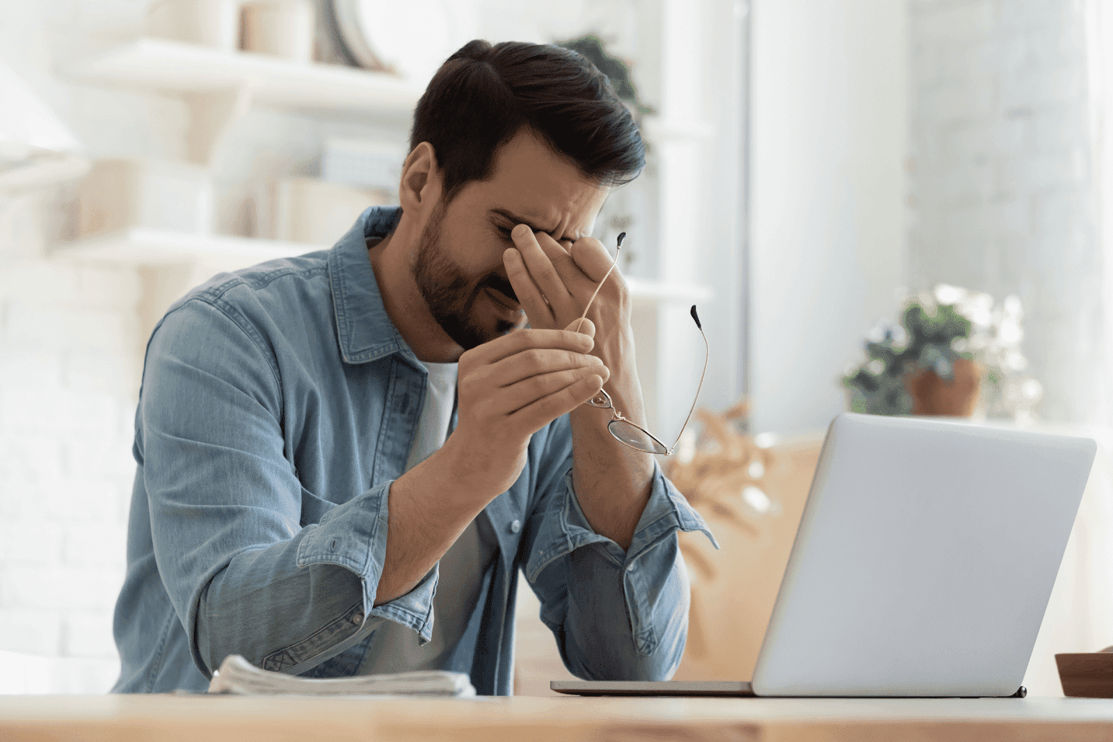 Man rubbing his eyes while holding glasses and sitting at a laptop