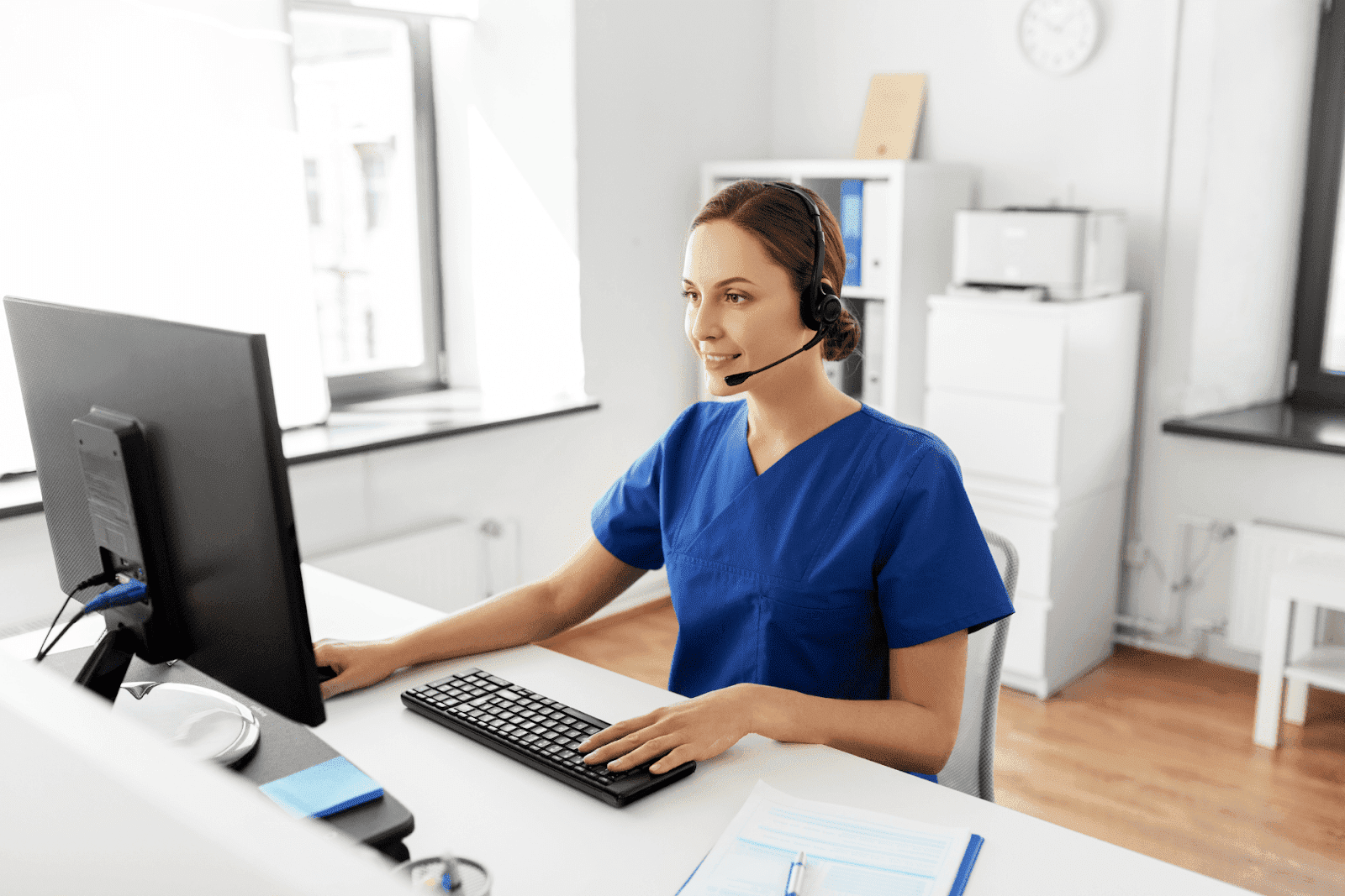 Medical professional in blue scrubs using a computer and wearing a headset during an online consultation